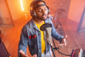 Portrait of a black young adult music artist recoding a rap song in front of the microphone and located in a small studio. He is wearing headphones and waving his hands. He is surrounded by recording equipment | edtechreader